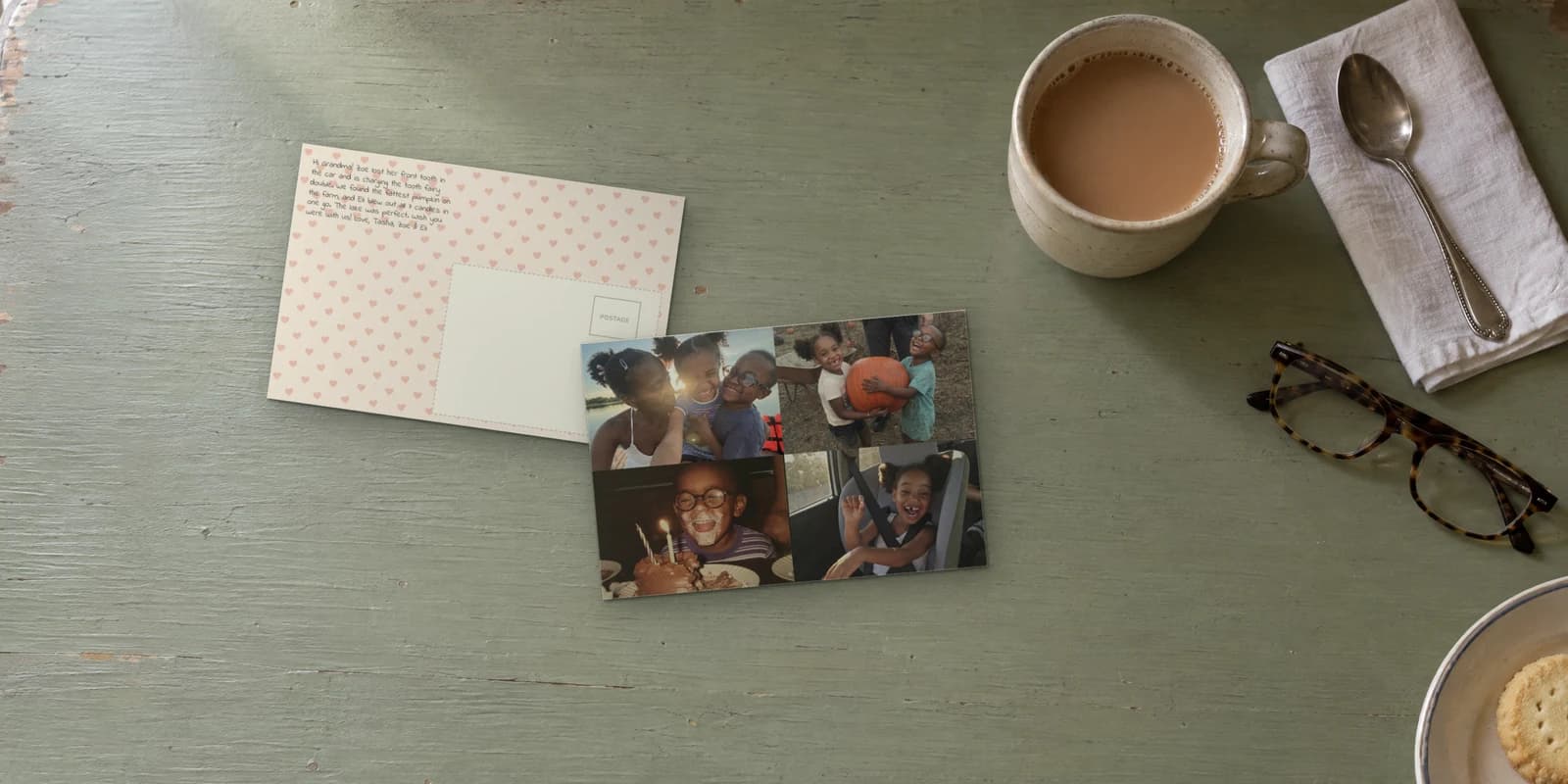 Send postcards like this one: a FamilyLetter 4x6 photo postcard with four photos of the grandkids, lying on Grandma's green kitchen table on top of its heart-patterned message side, next to a mug of tea, her reading glasses and a plate of cookies