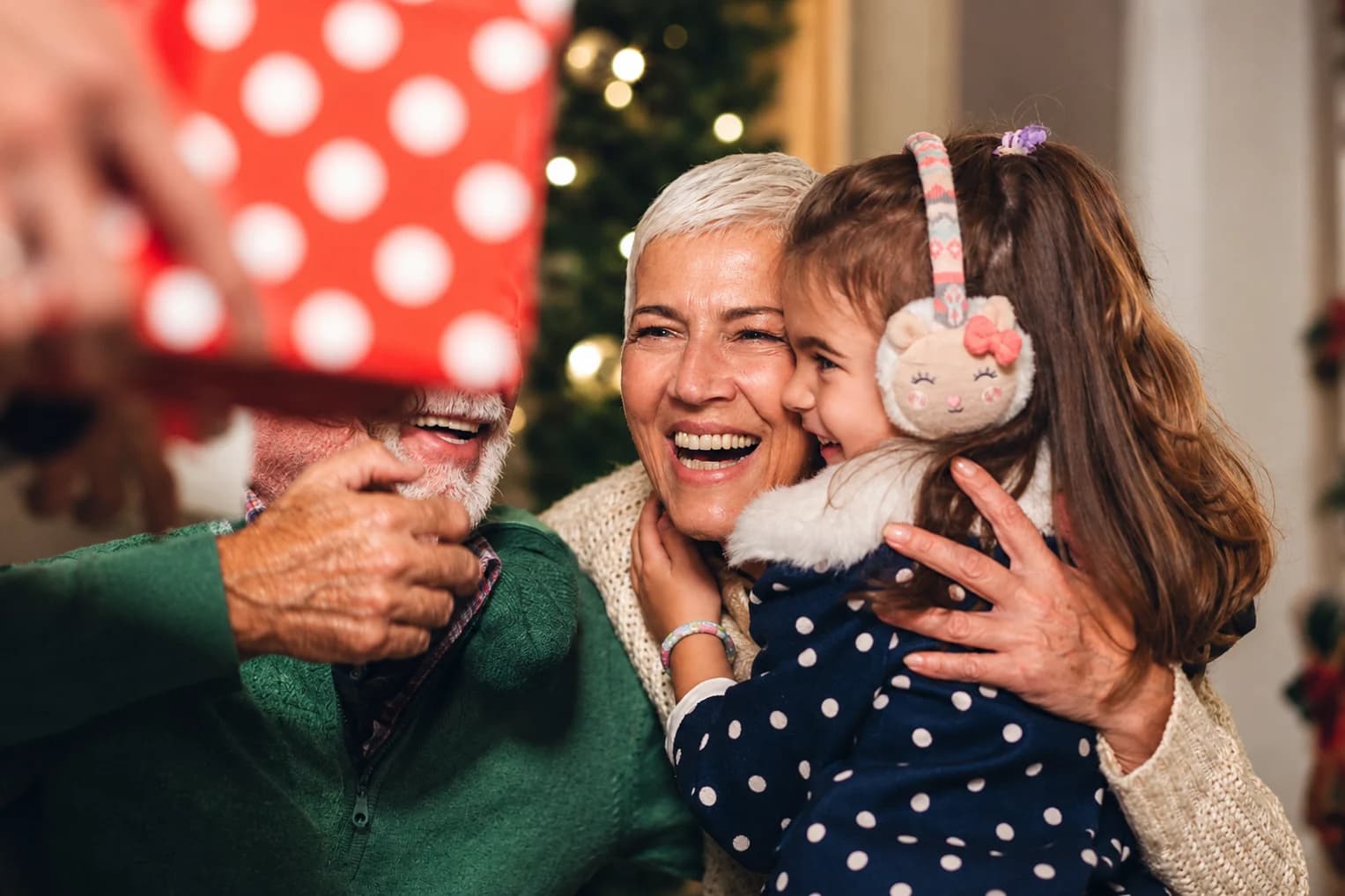 Christmas gifts for grandma: a grandmother laughing with her granddaughter in her arms beside the tree on Christmas morning
