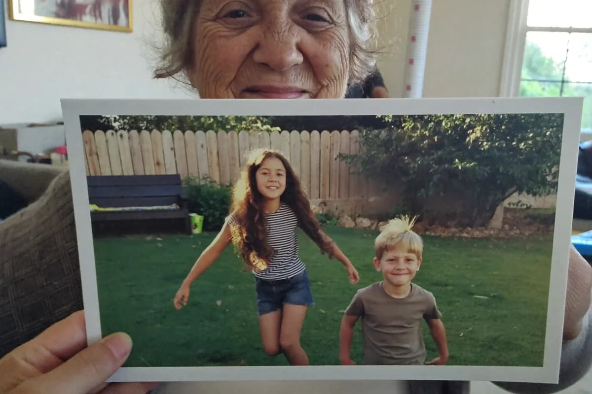 A grandmother holding up a printed 4×6 photo postcard showing two of her grandchildren in the garden