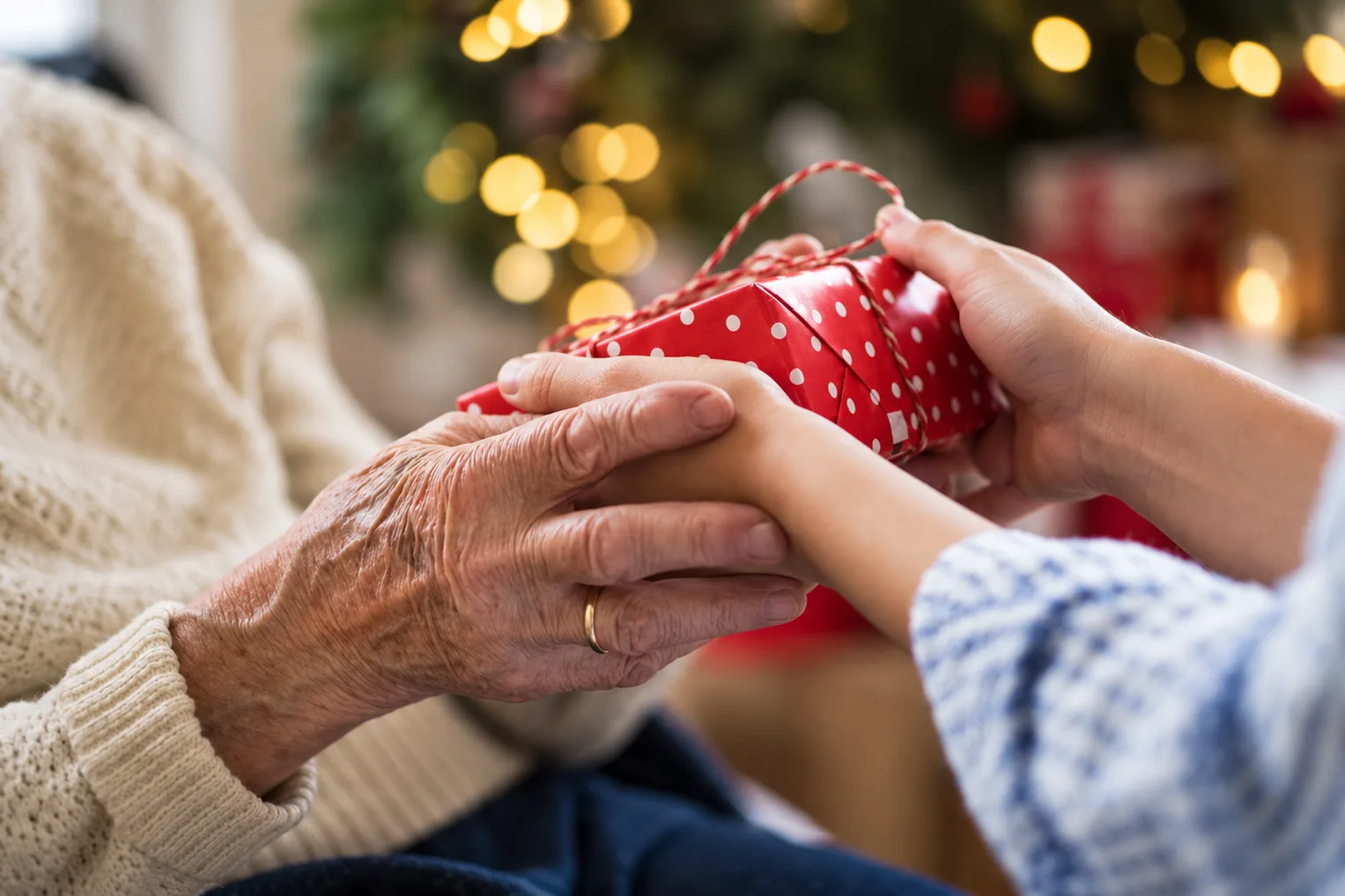 Christmas gifts for grandparents: an older hand wearing a wedding ring passing a small red wrapped present to a younger pair of hands beside a lit Christmas tree