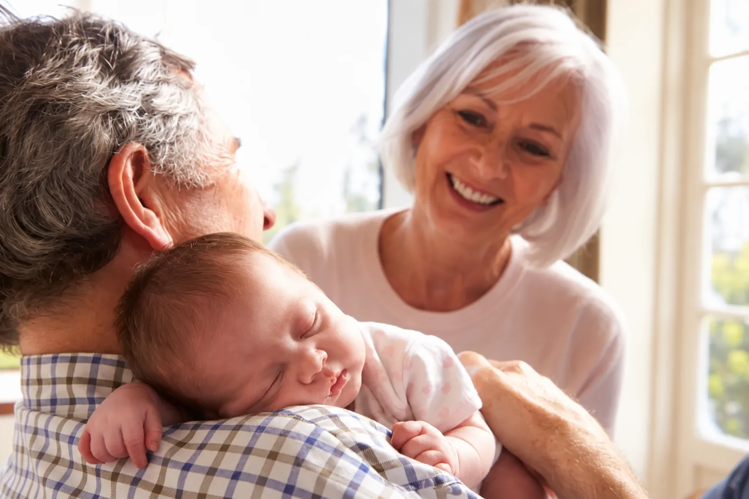 Gifts for new grandparents: a grandfather holding his sleeping newborn grandchild while the grandmother leans in, smiling