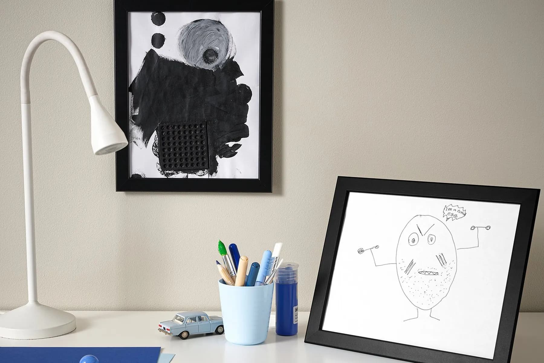 Homemade gifts for grandparents: two children's drawings in black frames, one hung on the wall and one standing on a desk beside a cup of pencils