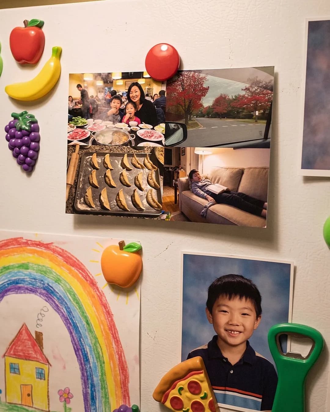 A photo postcard held on the fridge with fruit magnets, beside a school photo and a child's rainbow drawing