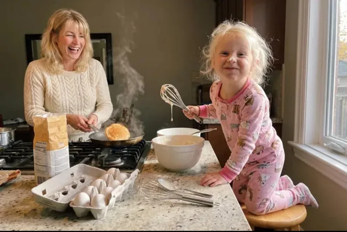 Card front with one photo of a grandmother laughing at the stove flipping a pancake while a toddler in pink pajamas holds up a whisk