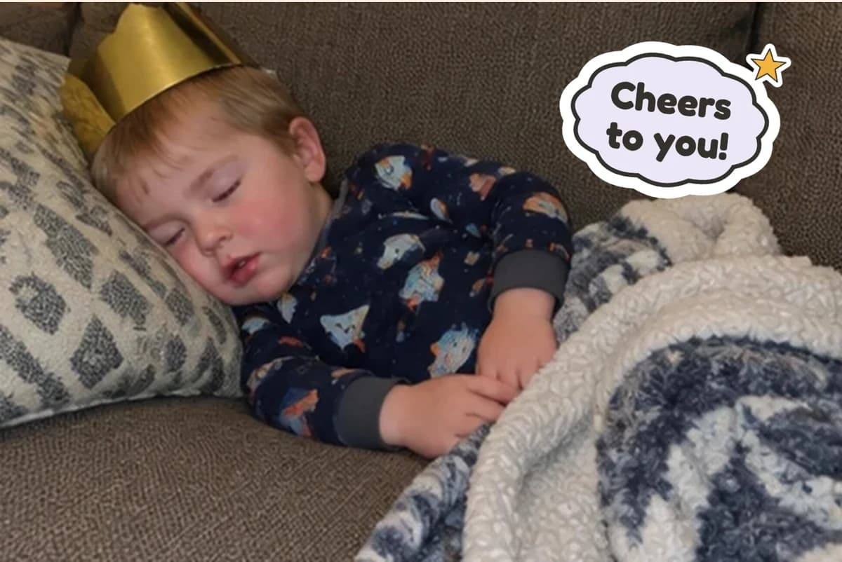 Card front with one photo of a toddler asleep on the couch in a gold paper crown and rocket pajamas, with a Cheers to you! sticker